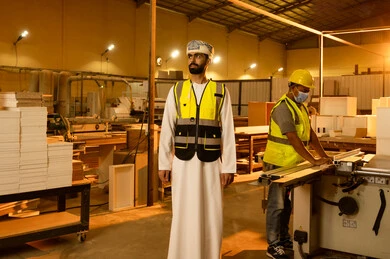 العمل في القطاع الصناعي، التطور والتقدم التقني في صناعة الخشب، معمل تصنيع الأثاث المنزلي، عامل يقوم بالعمل على جهاز قص الخشب بالليزر، مهندس عربي خليجي عماني يرتدي العمامة و ستره الحماية يقوم بالاشراف على العمال