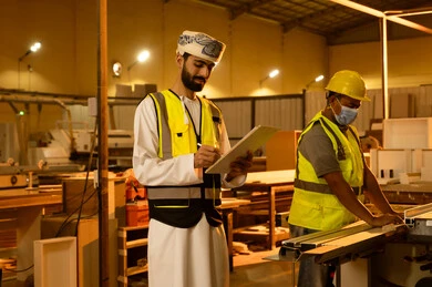تدوين الملاحظات الفنية، العمل في القطاع الصناعي، التطور والتقدم التقني في صناعة الخشب، معمل تصنيع الأثاث المنزلي، عامل يقوم بالعمل على جهاز قص الخشب بالليزر، مهندس عربي خليجي عماني يرتدي العمامة و ستره الحماية يقوم بالاشراف على العمال