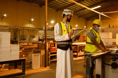 The development and technological advancement in the wood industry, a workshop for manufacturing home furniture, a worker operating a laser wood cutting machine, writing and taking notes, working in the industrial sector, an Arab Gulf Omani engineer wearing a turban and protective vest supervising the workers.