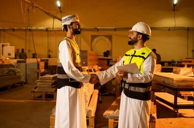 تبادل التحية والسلام، ورشات عمل في سلطنة عمان, جودة الانتاج المحلي الوطني, مهن وحرف شعبية تقليدية قديمة, صناعة الأثاث المنزلي والمكتبي، رجل مهندس عربي خليجي عماني يرتدي الثوب التقليدي وزي العمل يصافح زميله