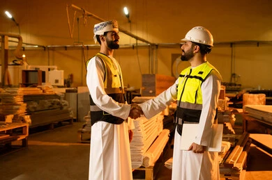 مصافحة ودية للترحيب، ورشات عمل في سلطنة عمان, جودة الانتاج المحلي الوطني, مهن وحرف شعبية تقليدية قديمة , صناعة الأثاث المنزلي والمكتبي، رجل مهندس عربي خليجي عماني يرتدي الثوب التقليدي وزي العمل يصافح زميله