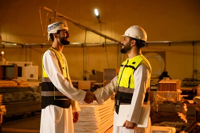 Greeting upon meeting, workshops in the Sultanate of Oman, quality of local national production, traditional old crafts and professions, manufacturing of home and office furniture, an Arab Gulf Omani engineer wearing traditional attire and work uniform shaking hands with his colleague.