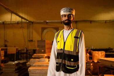 Design and manufacturing of home and office furniture, quality of local national production, an Arab Gulf Omani engineer wearing a dishdasha, turban, and safety vest looking at the camera, standing upright with expressions of joy, workshops in the Sultanate of Oman.