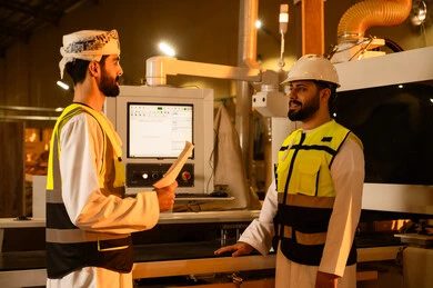 تشغيل الأجهزة عبر التقنيات الحديثة، المتابعة والإشراف على آلات المعمل، مصنع تصنيع الأثاث الخشبي، مهن ووظائف صناعية، ورشات النجارة في سلطنه عمان،مهندسان عربيان خليجيان عمانيان يرتديان سترة وخوذة الحماية يقفان داخل المصنع يقفان داخل المصنع، تبادل الافكار و الخبرات