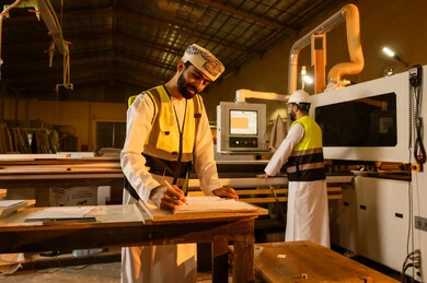 التطور والتقدم التقني في صناعة الخشب، عامل يقوم بالعمل على جهاز قص الخشب بالليزر، كتابة و تدوين الملاحظات، معمل تصنيع الأثاث المنزلي، العمل في القطاع الصناعي، مهندس عربي خليجي عماني يرتدي الدشداشة و ستره الحماية يقوم بالاشراف على العمال