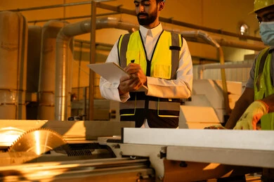 A worker operating a laser wood cutting machine, writing and taking notes, a home furniture manufacturing workshop, technological development and progress in the wood industry, working in the industrial sector, an Arab Gulf engineer in a white thobe and safety vest supervising the workers.