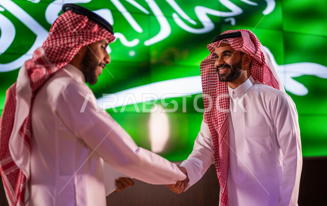 رجلان عربيان خليجيان سعوديان يتصافحان امام خلفية لعلم المملكة العربية السعودية ، لقاء وتعارف ، اتفاق عمل ،اليوم الوطني السعودي ، الاحتفال بعيد الاستقلال السعودي ، ذكرى تاسيس المملكة العربية السعودية
