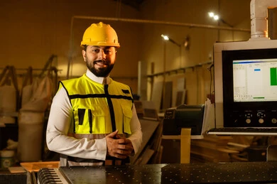 Factories and workshops in the Kingdom of Saudi Arabia, a wood manufacturing workshop, supervising the progress of the work plan, workshops and traditional crafts, a Saudi Gulf Arab engineer wearing a helmet and safety vest looking at the camera with expressions of happiness and joy.