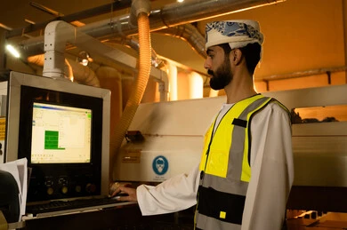 التحكم بالأجهزة من خلال التكنولوجيا،المتابعة والإشراف على آلات المعمل، مصنع تصنيع الأثاث الخشبي، مهن ووظائف صناعية، ورشات النجارة في سلطنه عمان، صورة  من الجانب لمهندس عربي خليجي عماني يرتدي العمامة وسترة الحماية يقف داخل المصنع