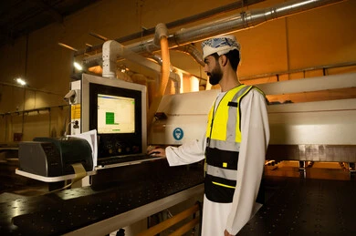 مراقبة الأجهزة أثناء التشغيل، التحكم بالمعدات باستخدام التقنية والتكنولوجيا، معمل تصنيع الأثاث الخشبي، مهن ووظائف صناعية، ورشات النجارة في سلطنه عمان، صورة  من الجانب لمهندس عربي خليجي عماني يرتدي العمامة وسترة الحماية يقف داخل المصنع