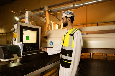 التحكم بالمعدات باستخدام التقنية والتكنولوجيا، معمل تصنيع الأثاث الخشبي، مهن ووظائف صناعية، ورشات النجارة في سلطنه عمان، المتابعة والإشراف على آلات المعمل، صورة  من الجانب لمهندس عربي خليجي عماني يرتدي العمامة وسترة الحماية يقف داخل المصنع