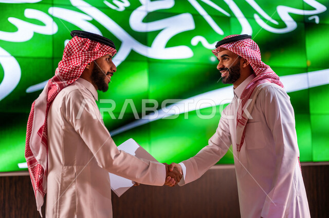 رجلان عربيان خليجيان سعوديان يتصافحان امام خلفية لعلم المملكة العربية السعودية ، لقاء وتعارف ، اتفاق عمل ،اليوم الوطني السعودي ، الاحتفال بعيد الاستقلال السعودي ، ذكرى تاسيس المملكة العربية السعودية