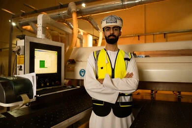 النظر إلى الكاميرا بإيماءات الثقة بالنفس والجدية، الاشراف على سير خطة العمل, ورشات عمل ومهن تقليدية قديمة, مصانع ومعامل في سلطنة عمان, مهندس عربي خليجي عماني يرتدي العمامة وسترة الحماية يقف بايدي متقاطعة، معمل صناعة الخشب