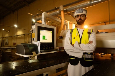 الاشراف على سير خطة العمل، النظر إلى الكاميرا بإيماءات الثقة بالنفس والجدية, ورشات عمل ومهن تقليدية قديمة, مصانع ومعامل في سلطنة عمان, مهندس عربي خليجي عماني يرتدي العمامة وسترة الحماية يقف بايدي متقاطعة، معمل صناعة الخشب