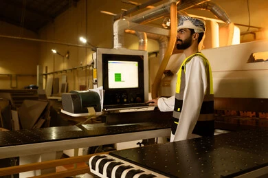 Controlling equipment using technology and techniques, monitoring the performance of industrial devices, wooden furniture manufacturing workshop, industrial professions and jobs, carpentry workshops in the Sultanate of Oman, a side view of an Arab Gulf Omani engineer wearing a turban and safety vest standing inside the factory.