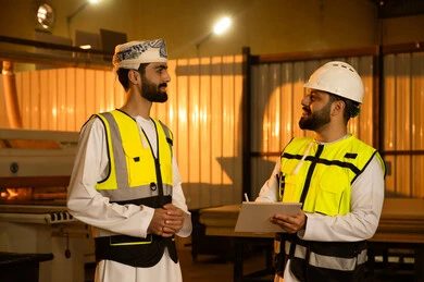 Sharing experiences and ideas, manufacturing home and office furniture, workshops in the Sultanate of Oman, quality of national local production, an Arab Gulf Omani engineer wearing a dishdasha, turban, and safety vest stands alongside a colleague as they monitor the workflow, writing and taking notes.