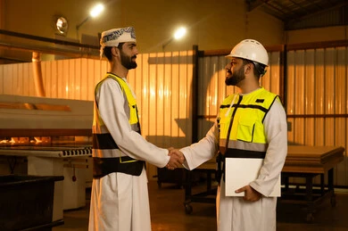 Welcoming colleagues in the workplace, workshops in the Sultanate of Oman, quality of local national production, traditional old crafts and professions, manufacturing of home and office furniture, an Arab Gulf Omani engineer wearing a dishdasha and turban and safety vest shaking hands with his colleague.