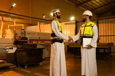 تبادل التحية مع الزملاء، ورشات عمل في سلطنه عمان, جودة الانتاج المحلي الوطني, مهن وحرف شعبية تقليدية قديمة , صناعة الأثاث المنزلي والمكتبي، رجل مهندس عربي خليجي عماني يرتدي الدشداشة و العمامة و ستره الحماية يقوم بمصافحة زميله