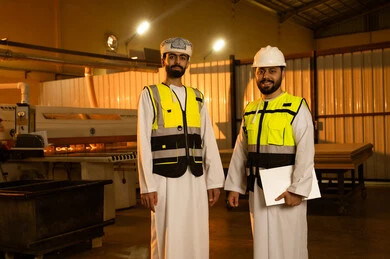 Industrial professions and jobs, wooden furniture manufacturing workshop, monitoring and supervising the workshop machines, carpentry workshops in the Sultanate of Oman, two Arab Gulf engineers from Oman wearing vests and safety helmets standing inside the factory looking at the camera with expressions of joy.