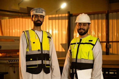 معمل تصنيع الأثاث الخشبي، مهن ووظائف صناعية، المتابعة والإشراف على آلات المعمل، ورشات النجارة في سلطنة عمان، مهندسان عربيان خليجيان عمانيان يرتديان سترة وخوذة الحماية يقفان داخل المصنع ينظران الى الكاميرا بتعابير السرور