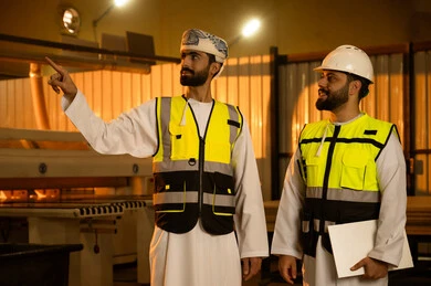 فحص ومراقبة معدات المعمل ، الاشارة الى شيء ما، ورشات النجارة في سلطنة عمان، معمل تصنيع الأثاث الخشبي، مهن ووظائف صناعية، مهندس عربي خليجي عماني يرتدي العمامة و سترة الحماية يقوم بمناقشة العمل مع زميلة داخل المصنع