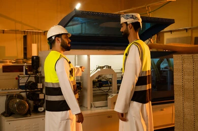 Supervising the operation of laboratory equipment, pointing to something, carpentry workshops in the Sultanate of Oman, wooden furniture manufacturing factory, industrial professions and jobs, an Omani Arab engineer wearing a turban and safety vest discussing work with a colleague inside the factory.