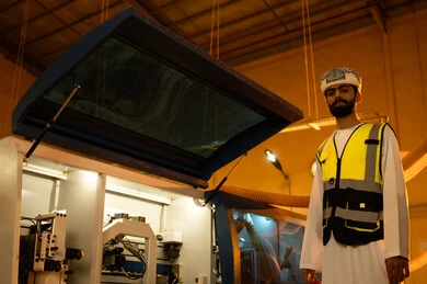 Supervising the implementation of planned tasks, quality of national local production, manufacturing of home and office furniture, an Arab Gulf Omani engineer wearing a dishdasha, turban, and safety vest looking at the camera, standing upright with expressions of joy, workshops in the Sultanate of Oman.