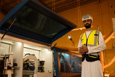 Monitoring the progress of work stages, the quality of national local production, the manufacture of home and office furniture, an Arab Gulf Omani engineer wearing a dishdasha, turban, and safety vest looking at the camera, standing with hands intertwined with expressions of joy, workshops in the Sultanate of Oman.
