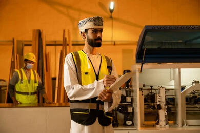 Wood furniture shaping workshop, industrial professions and jobs, recording and taking notes, monitoring and supervising laboratory machines, carpentry workshops in the Sultanate of Oman, an Arab Gulf Omani engineer wearing a turban and dishdasha and a safety vest holding a paper folder in his hand.