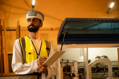 Wooden furniture manufacturing factory, industrial professions and jobs, recording and taking notes, monitoring and supervising workshop machines, carpentry workshops in the Sultanate of Oman, an Arab Gulf engineer from Oman wearing a turban, dishdasha, and safety vest holding a paper folder in his hand.