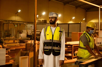 إنشاء وتصنيع أثاث المنازل والمكاتب، جودة الانتاج المحلي الوطني، رجل مهندس عربي خليجي عماني يرتديالدشداشة و العمامة و سترة الحماية ينظر الى شيء ما ، الوقوف باستقامة بتعابير السرور، ورشات عمل في سلطنة عمان