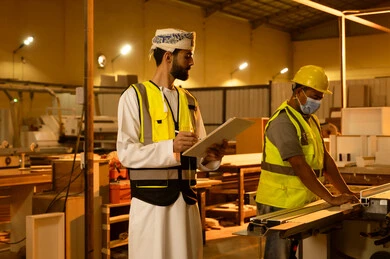 كتابة و تدوين الملاحظات، عامل يقوم بالعمل على جهاز قص الخشب بالليزر، معمل تصنيع الأثاث المنزلي، التطور والتقدم التقني في صناعة الخشب، العمل في القطاع الصناعي، مهندس عربي خليجي عماني يرتدي الدشداشة و ستره الحماية يقوم بالاشراف على العمال