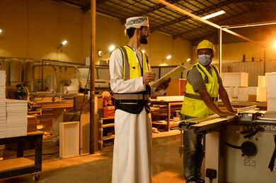 العمل في القطاع الصناعي، التطور والتقدم التقني في صناعة الخشب، مهندس عربي خليجي عماني يرتدي الدشداشة و ستره الحماية يقوم بالاشراف على العمال، معمل تصنيع الأثاث المنزلي، عامل يقوم بالعمل على جهاز قص الخشب بالليزر، كتابة و تدووين الملاحظات