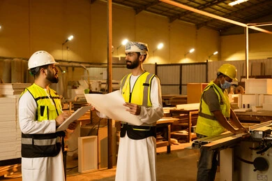 The quality of national local production, the manufacture of home and office furniture, workshops in the Sultanate of Oman, exchanging opinions and experiences, an Arab Gulf Omani engineer wearing traditional attire and a safety vest holding the engineering plan and discussing with his colleague inside the factory.
