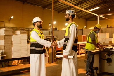 Peace and greeting exchange, workshops in the Sultanate of Oman, quality of local national production, traditional old crafts and professions, manufacturing of home and office furniture, an Arab Gulf Omani engineer wearing a dishdasha, turban, and protective vest shakes hands with his colleague.