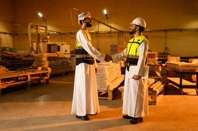 إلقاء التحية والمصافحة، ورشات عمل في سلطنه عمان, جودة الانتاج المحلي الوطني, مهن وحرف شعبية تقليدية قديمة , صناعة الأثاث المنزلي والمكتبي، رجل مهندس عربي خليجي عماني يرتدي الدشداشة و العمامة و ستره الحماية  يقوم بمصافحة زميله