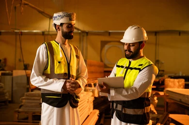 The concept of discussions and dialogues, note-taking through writing, a wooden furniture manufacturing workshop, two Arab Gulf engineers from Oman wearing vests and safety helmets supervising the progress of the work plan, popular crafts and manual professions, carpentry workshops in the Sultanate of Oman.