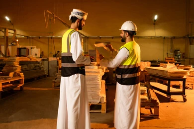 The development and technological advancement in the wood industry, monitoring and supervising the workshop machines, working in the industrial sector, two Arab Gulf engineers from Oman wearing helmets and safety vests working in a carpentry workshop, recording and noting observations, home furniture manufacturing workshop.