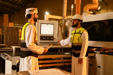Exchange of opinions and experiences, monitoring and supervising laboratory machines, wooden furniture manufacturing workshop, industrial professions and jobs, controlling equipment using technology and techniques, an Arab Gulf Omani engineer wearing a turban and protective jacket stands with a colleague holding a bundle of paper in his hands.