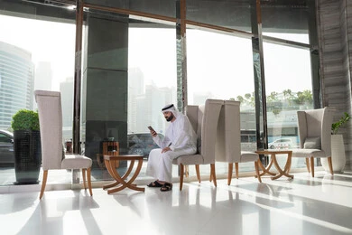 تصفح تطبيقات الموبايل، رجل عربي خليجي اماراتي يرتدي الكندورة والغترة يجلس داخل الفندق يقوم بالدردشة مع الاهل والاصدقاء بايماءات السرور والسعادة، متابعة مواقع التواصل الاجتماعي عبر الهاتف المحمول، دمج التقنيات الحديثة بالحياة العملية