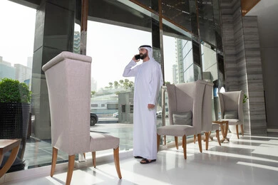 Gestures of happiness in communicating with friends and relatives, enjoying exchanging conversations and news, making a voice phone call via mobile, an Emirati Arab man wearing the kandura and Emirati ghutrah standing inside the hotel talking on his mobile phone.