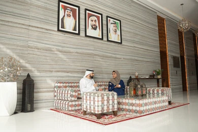 Discussion and exchange of information, strengthening marital relationships and bonds of love and affection, enjoying hot beverages, the concept of a happy family, an Emirati Arab couple wearing traditional attire sitting in a ground-level popular gathering, gestures of happiness and joy.