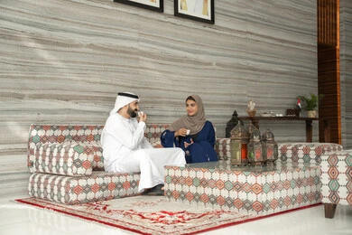 Strengthening marital relationships and bonds of love and affection, exchanging news and family dialogues, enjoying hot beverages, the concept of a happy family, an Emirati Arab couple wearing traditional attire sitting in a ground-level popular gathering, gestures of happiness and joy.