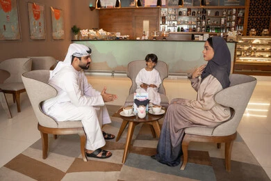 The concept of a happy family, exchanging conversations and news with gestures of joy and happiness, an Emirati Arab couple wearing traditional attire sitting in a café with their son, socializing and building relationships, enjoying hot beverages.