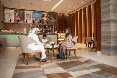 تبادل الأحاديث والأخبار بإيماءات السرور والفرح، زوجان عربيان خليجيان اماراتيان يرتديان الزي التقليدي يجلسان في كافية برفقة ابنهم , مفهوم الاسره السعيده, تعارف وبناء علاقات اجتماعيه, الاستمتاع بتناول المشروبات الساخنه