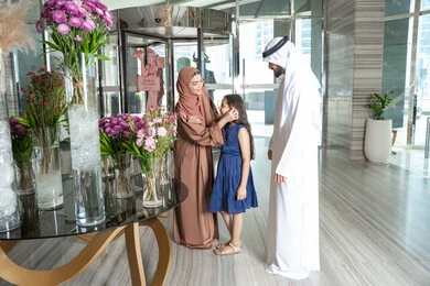Spending happy times with family, parents following up on their children and taking care of them, gestures of happiness and joy, an Emirati Gulf Arab mother wearing an abaya and hijab stands with her husband at the hotel reception entrance, placing a flower in her daughter's hair, preparing to enjoy a pleasant family trip.