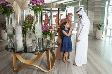 مشاعر الأمان والحب مع العائلة، ايماءات السعادة و السرور، رجل عربي خليجي اماراتي يرتدي الكندورة و الغتره يقف برفقة زوجته فى مدخل اسقبال الفندق يقوم بتقديم وردة الي ابنته، الاستعداد لقضاء رحلة سياحية ممتعه مع العائلة