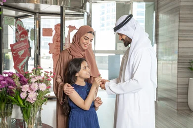 The concept of family bonding, gestures of happiness and joy, an Emirati Gulf Arab man wearing a kandura and ghutrah stands with his wife at the hotel reception entrance, presenting a flower to his daughter, preparing to enjoy a pleasant family trip.