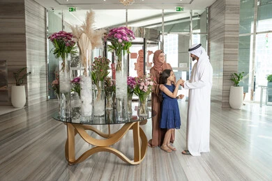 المحافظة على الترابط الأسري، ايماءات السعادة و السرور، رجل عربي خليجي اماراتي يرتدي الكندورة و الغتره يقف برفقة زوجته فى مدخل اسقبال الفندق يقوم بتقديم وردة الي ابنته، الاستعداد لقضاء رحلة سياحية ممتعه مع العائلة