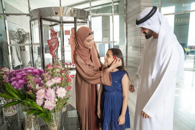 دور الآباء في رعاية أطفالهم، قضاء أوقات سعيدة برفقة الأهل، ايماءات السعادة و السرور، ام عربيه خليجيه اماراتية ترتدي العباءة و الحجاب تقف برفقة زوجها داخل مدخل استقبال الفندق تقوم بوضع وردة فى شعر ابنتها، الاستعداد لقضاء رحلة سياحية ممتعه مع العائلة